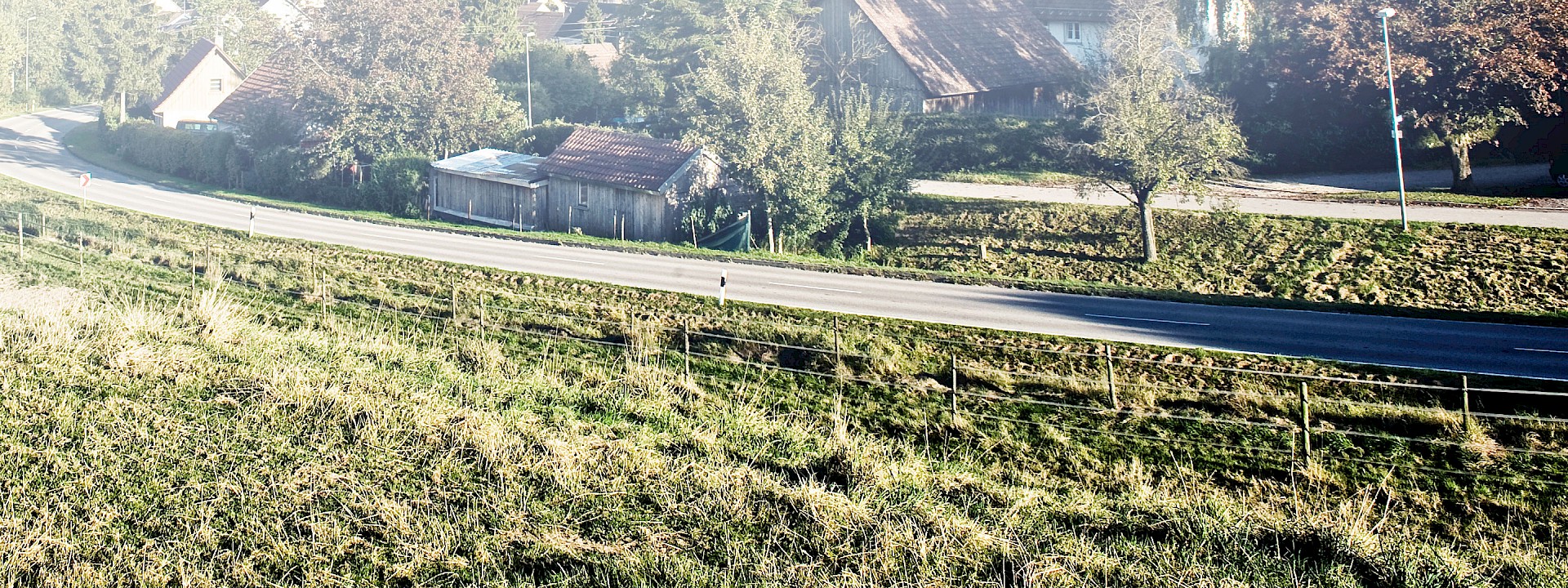 Straße an einem Weidehang und unterhalb der Straße ein alter Bauernhof