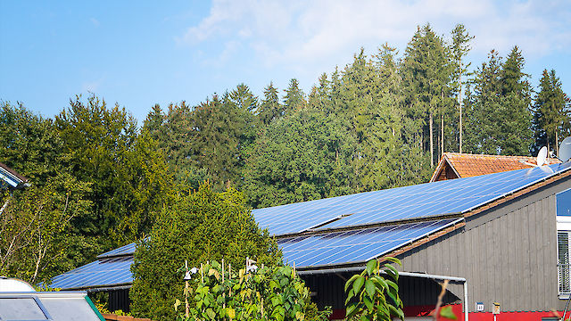 Gebäude mit PV-Analge auf dem Dach, im Vordergrund ein grüner Garten und im Hintergrund Wald