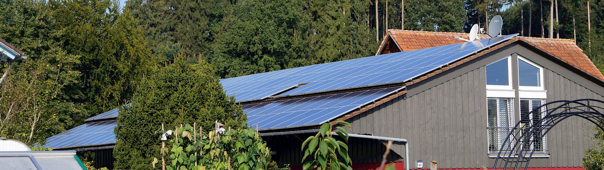 Gebäude mit PV-Analge auf dem Dach, im Vordergrund ein grüner Garten und im Hintergrund Wald