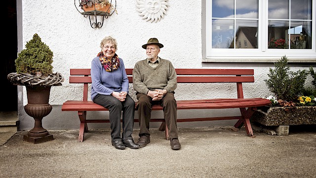 alte Frau und Mann auf einer Gartenbank vor einem Haus