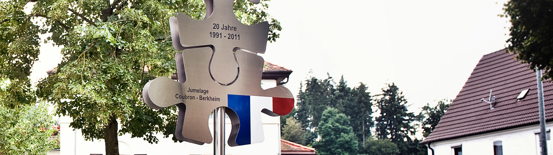 Schild aus Metall in Form eines Puzzels mit der Französischen Nationalflagge und dem Namen der Partnerstadt Coubron